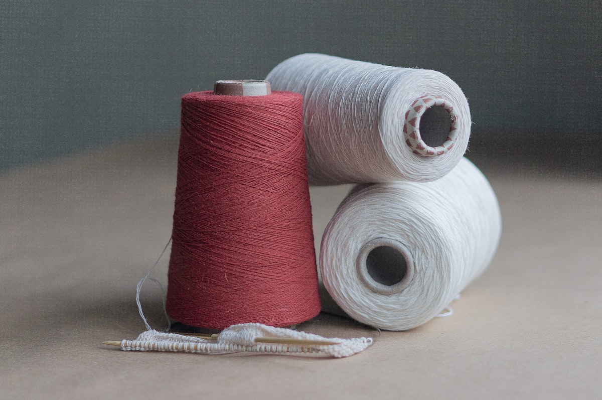 A still life of one deep red and two white cones of thread on a plain neutral surface, with a small piece of knitting on wooden needles in front, a neutral abstract garment-making image; no identifiable person and no face, no readable brand name or logo, no recognizable branded product, and it does not depict a sweatshop scene, for the investigation into Carabinieri raids on luxury houses over sweatshop labor in Italian supply chains