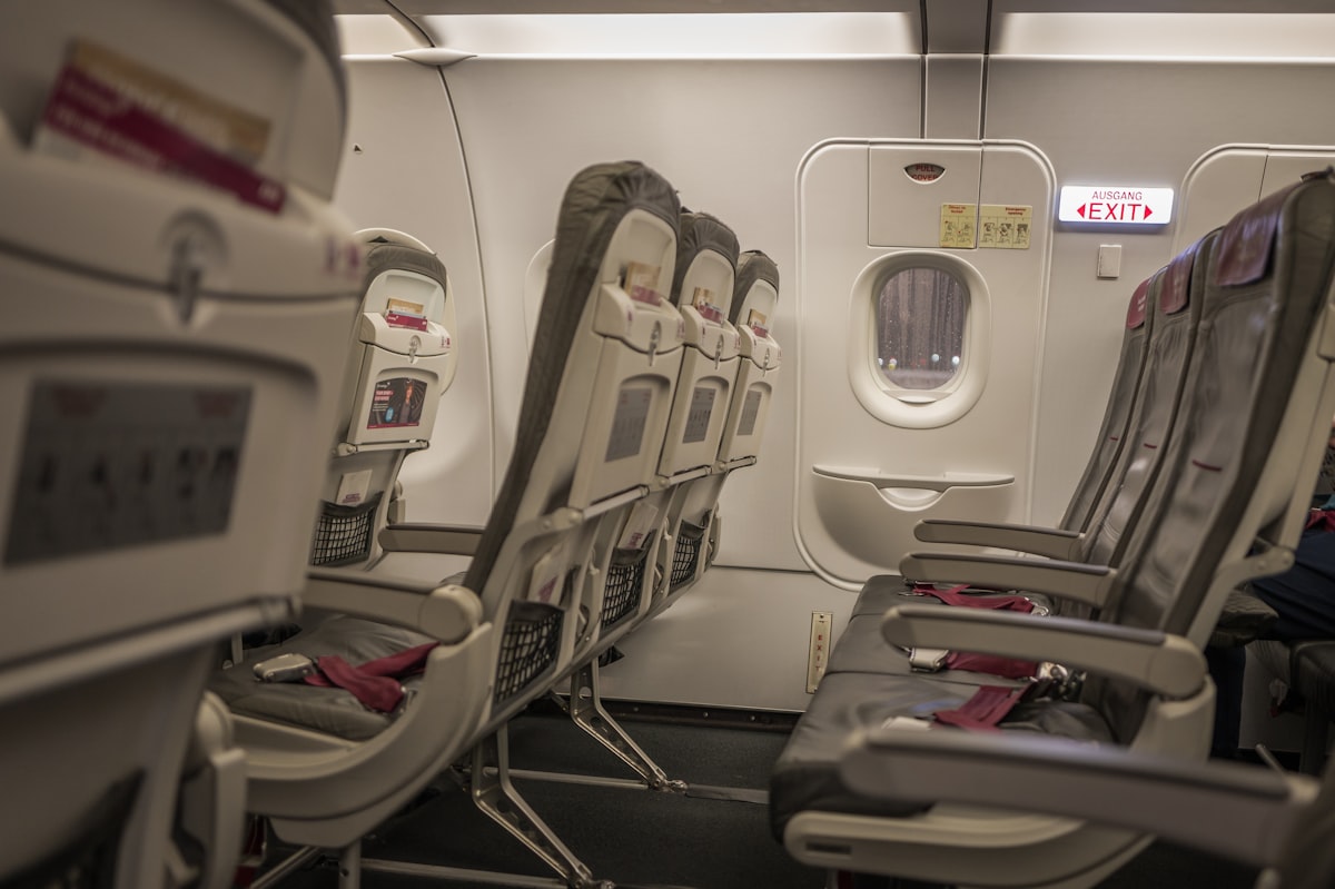 An empty economy-class cabin with rows of gray seats beside an exit-row window and a generic AUSGANG EXIT sign, no identifiable people and no readable airline brand or logo, for the piece on shrinking seat pitch and reclining etiquette