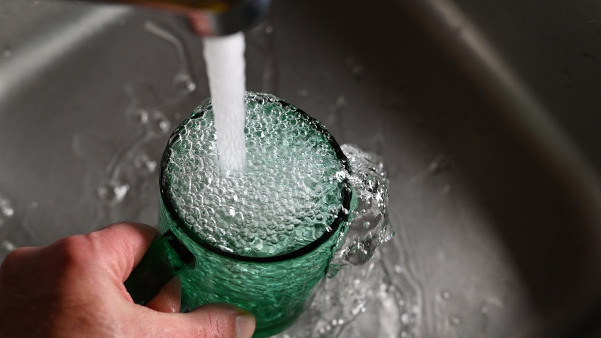 A hand holding a green glass under a running tap as it fills with water in a steel sink, no identifiable face and no readable brand or logo, for the piece on hydration science replacing the eight-glasses rule