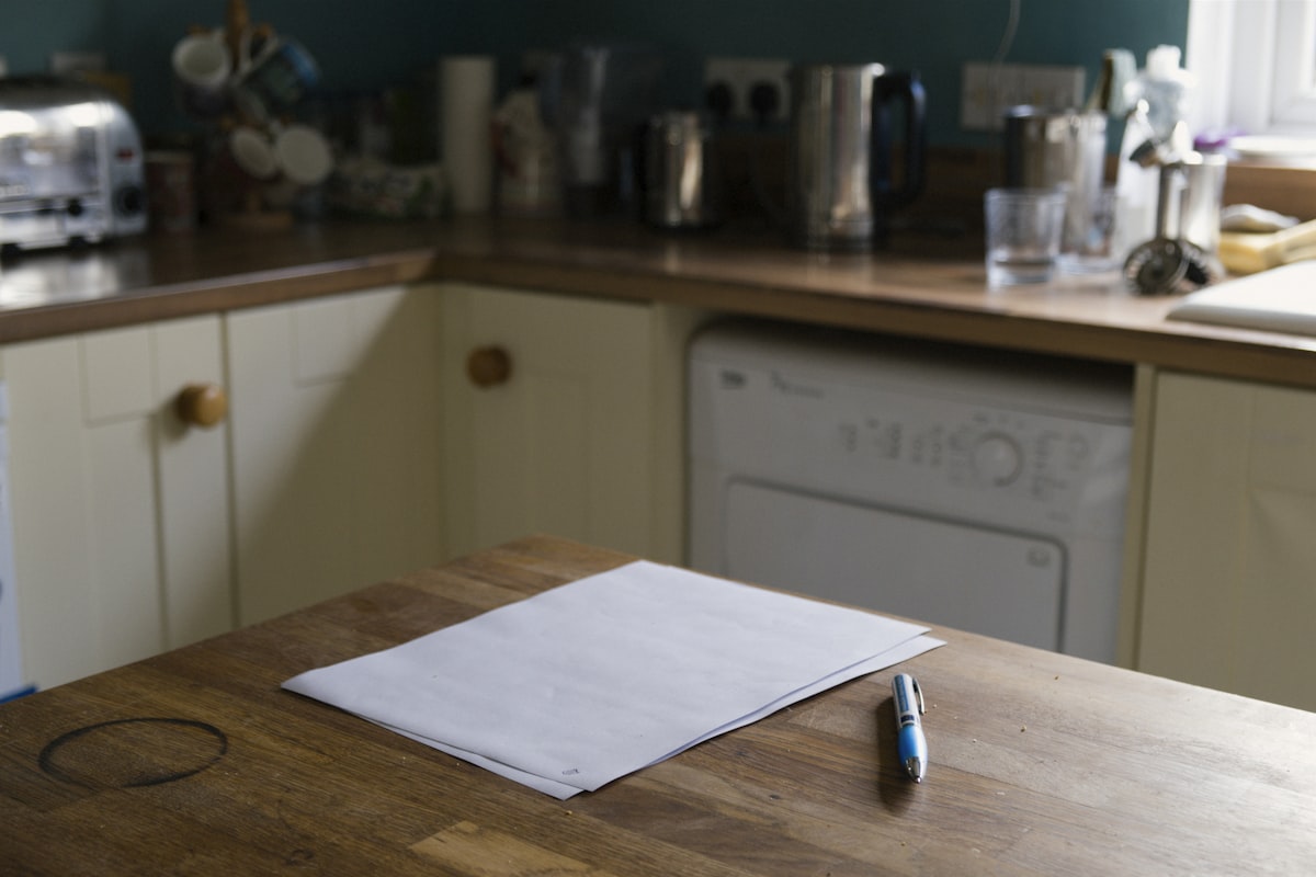A blank sheet of paper and a pen on a worn wooden kitchen table in a lived-in home kitchen, appliances softly out of focus behind, no identifiable person and no readable brand or logo, a quiet domestic still that reads as the household paperwork waiting to be dealt with, for the piece on the sandwich generation and the private cost of parent care