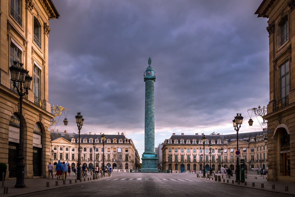 The Colonne Vendome rising from a near-empty Place Vendome at dusk, flanked by the square's classical facades, no identifiable people and no readable brand or logo, placing the piece at the address of Charvet's shop, for the piece on Chanel's acquisition of the shirtmaker Charvet