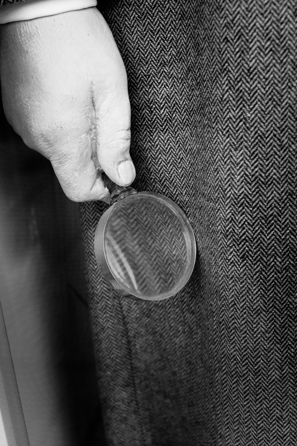 A close black-and-white still of a hand holding a round magnifying loupe against plain herringbone tweed, reading the weave up close, no identifiable person (a hand only, no face) and no readable brand, logo, monogram, or check pattern, for the piece on super-fakes outrunning the authenticator's loupe