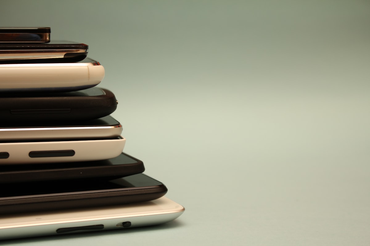 A stack of assorted switched-off smartphones piled on a plain neutral surface, no identifiable people and no readable brand or logo, evoking the phones-in-a-pile dinner ritual, for the piece on restaurants that lock diners' phones away