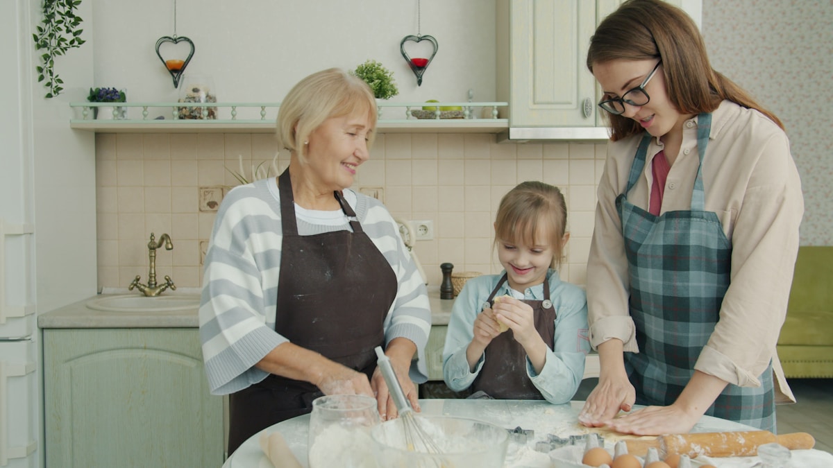 A grandmother, mother, and daughter baking together in a kitchen, three generations sharing an ordinary domestic task, for the piece on multigenerational households and the housing economics behind them