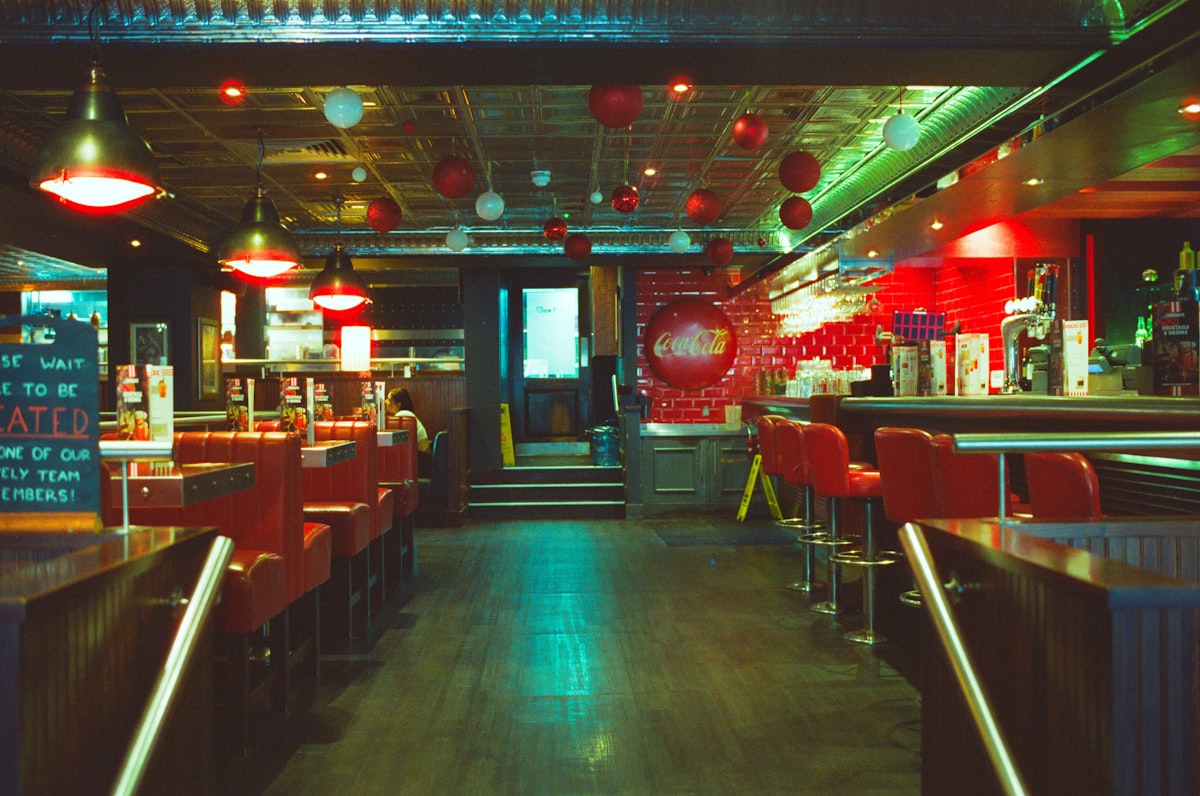 An empty retro diner at dusk, red bar stools along a counter and bare tables, no identifiable person and no readable brand or logo, for the piece on third places (the diner, the bar, the lodge) quietly closing, hardest where they were already scarce