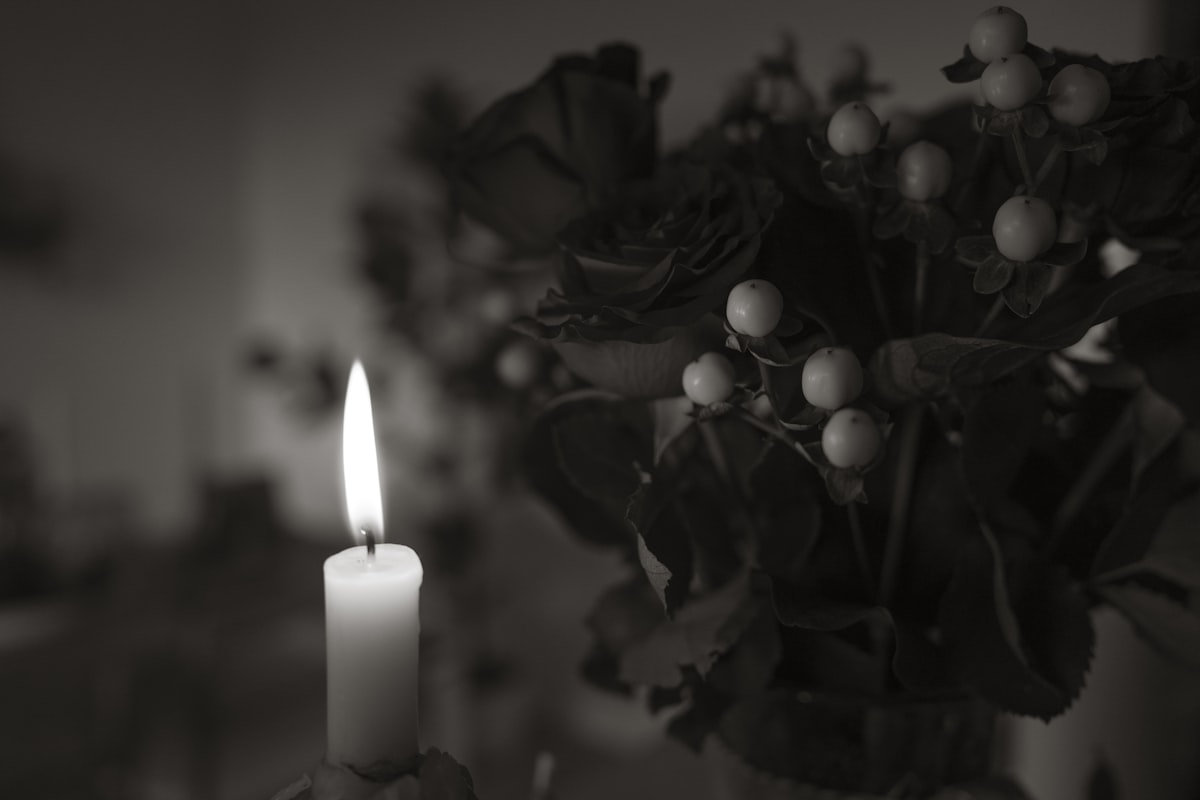 A single lit candle beside a bouquet of dark roses, no people, no identifiable person or place, for the piece on the collision between private mourning and public tribute online