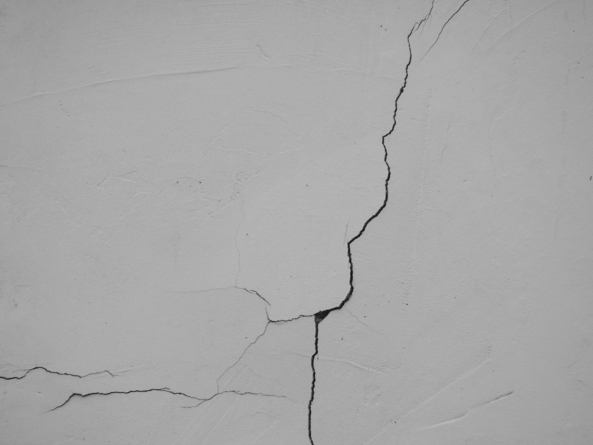 A hairline crack in an otherwise pristine white wall, no people, a literal visual for the piece on quiet cracking and the workers holding steady on the surface while coming apart underneath