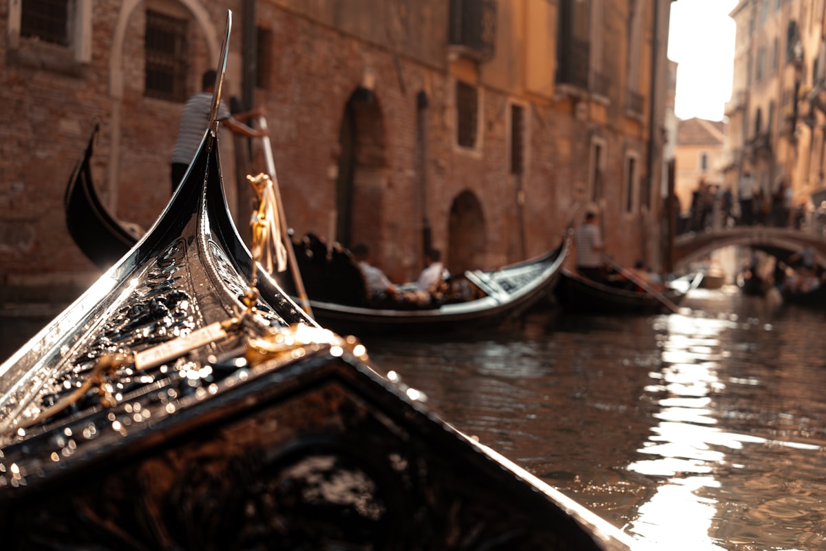 Gondolas cruising along a narrow Venice canal in warm light, the passengers distant and not individually identifiable, no readable brand or logo, for the piece on Italy turning tourist manners into cash penalties