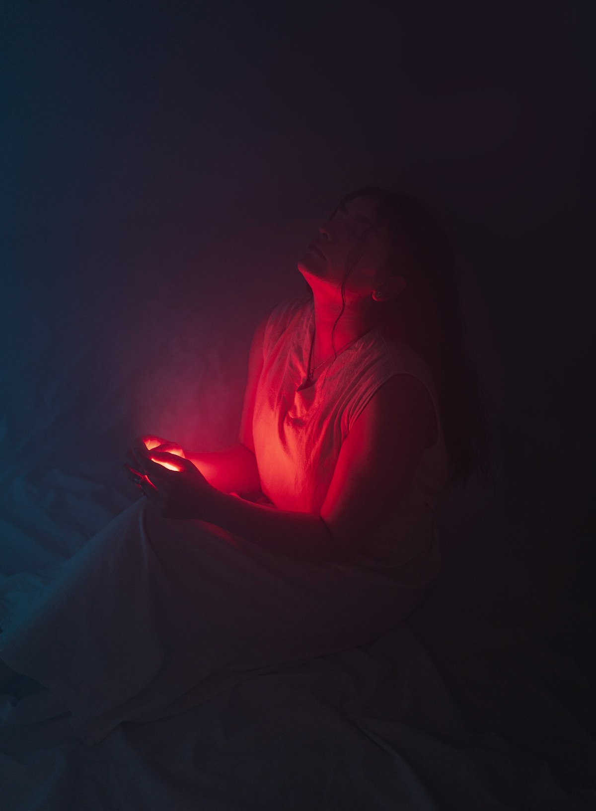 A person sitting in the dark, face obscured, lit only by the red glow of a phone screen held in both hands, for the piece on AI companion apps and the loneliness paradox they create