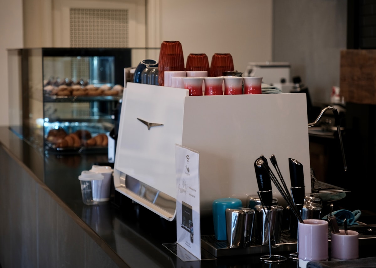 An espresso machine on a cafe counter with stacked cups and pastries in the soft-focus background, no identifiable person and no readable brand name or logo, for the piece on specialty coffee retreating from pour-over ritual toward simple, well-made cups