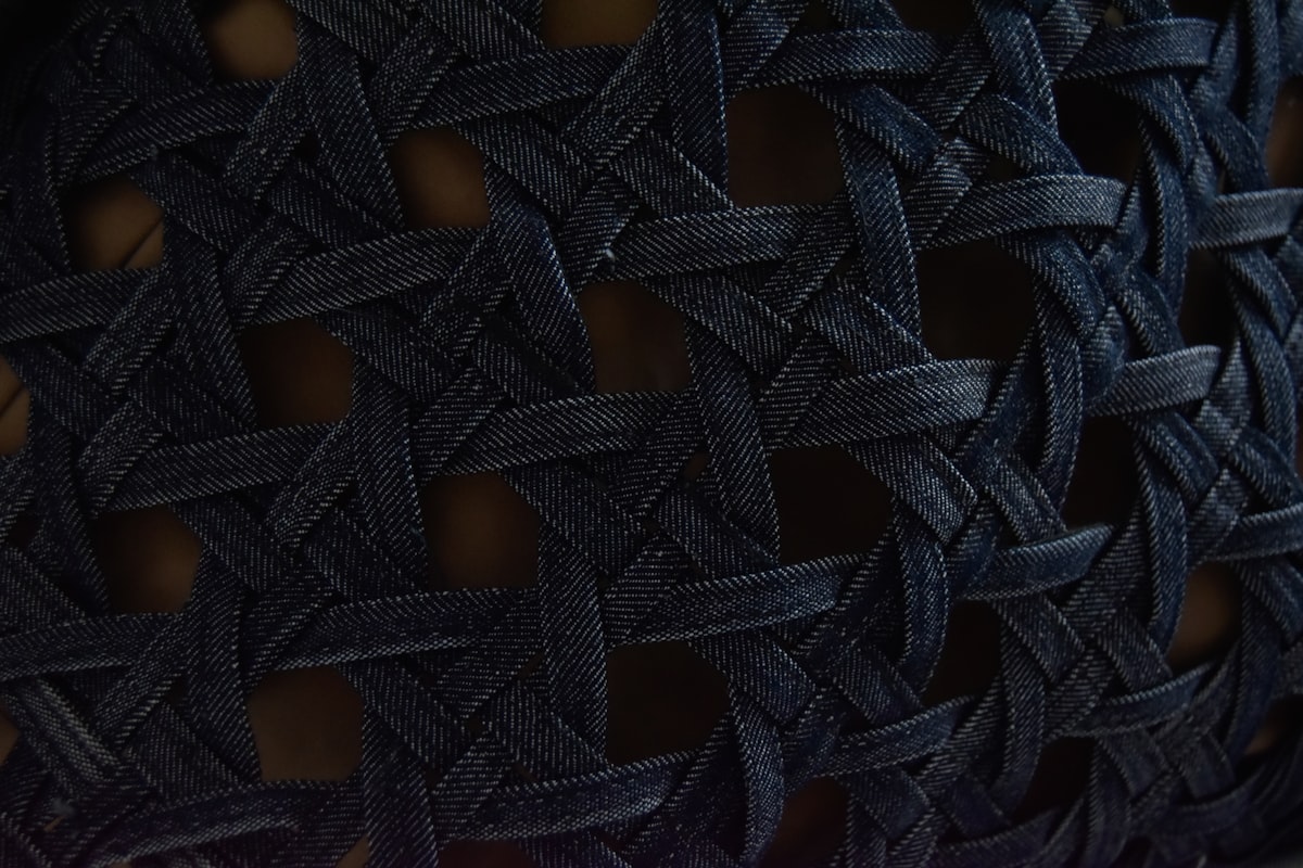 A tight close-up of a dark woven lattice, its strands crossing in a repeating intrecciato-style basketweave, no identifiable person and no readable brand or logo, standing in for the craft signal the reader is asked to learn to recognize, for the piece on luxury's shift from monograms to craft signals