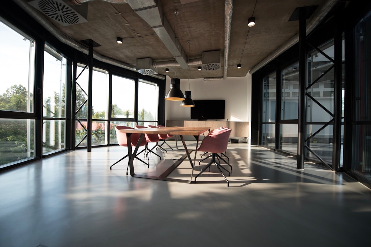A clean, empty modern meeting room with a long boardroom table and chairs beside a wall of windows, no identifiable person and no readable brand or logo, its vacant corporate setting framing the debate over who sets the rules of the room, for the piece on office etiquette and the cameras-on flashpoint