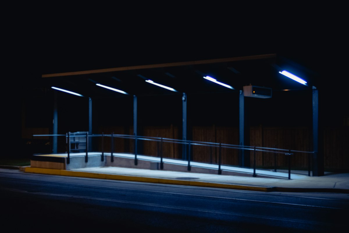 An empty, lit bus shelter beside a dark road at night; generic, not Western Sydney International or Sydney; no people, no vehicles, no airline, operator or transport-agency branding